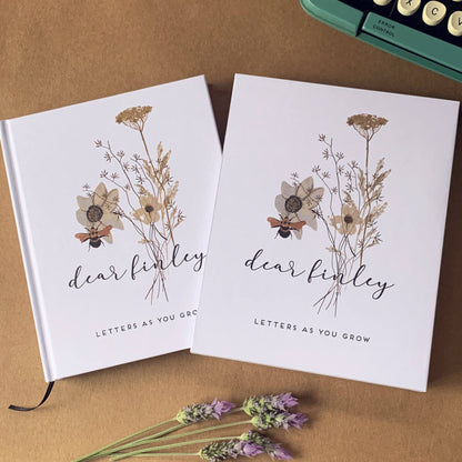 Custom pregnancy journal with hardback white cover, customized with a wildflower design and baby's name and title "Letters As You Grow". Housed in matching book slip case. On a table by old typewriter and lavender flowers.