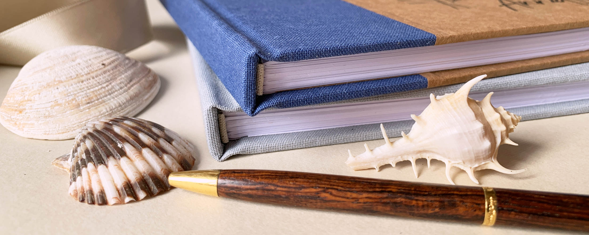 Two linen covered guest books, a pen, and seashells on a textured surface