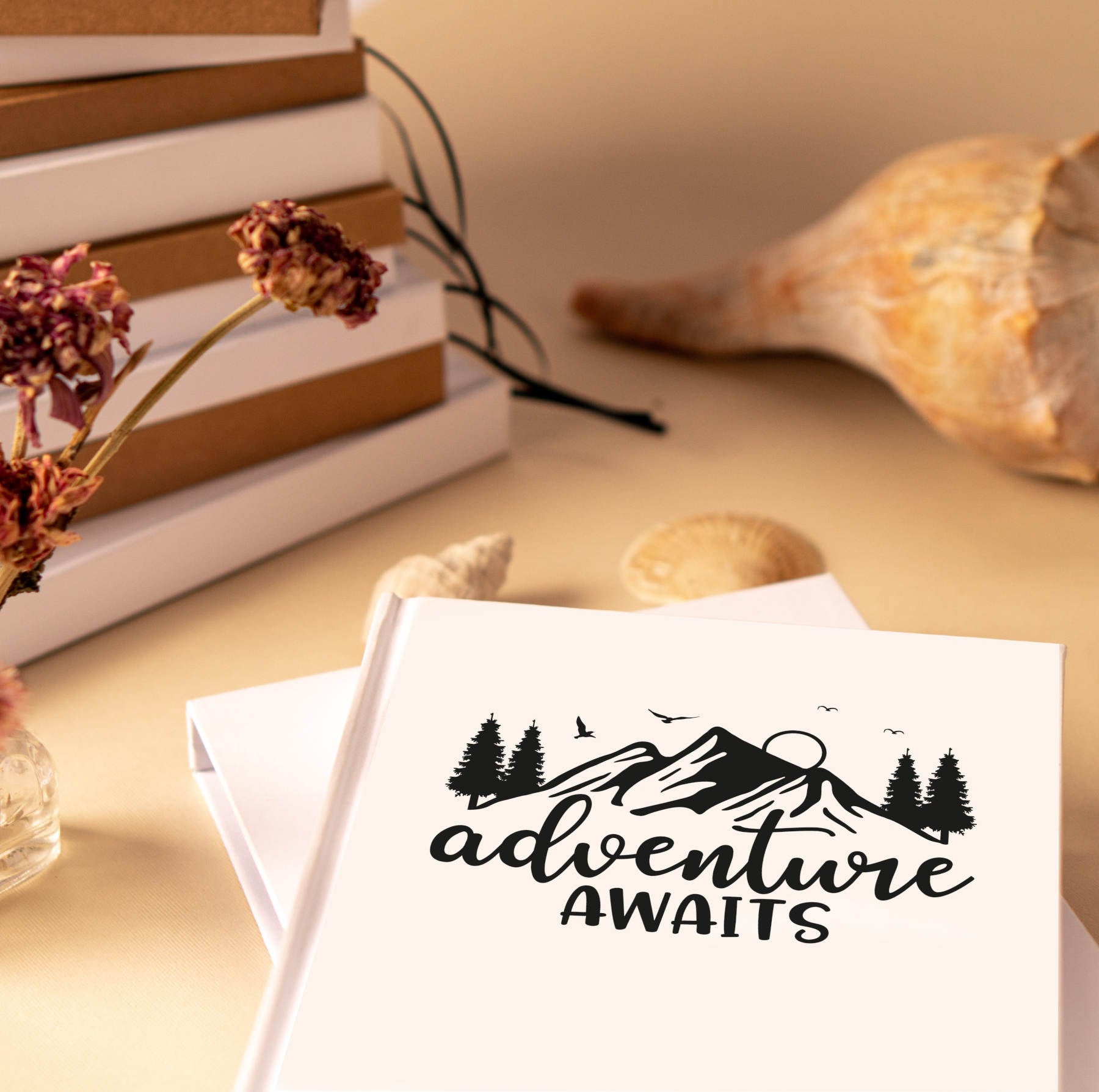White "Adventure Awaits" travel journal on a table top with shells, dried flowers, and a stack of books behind.