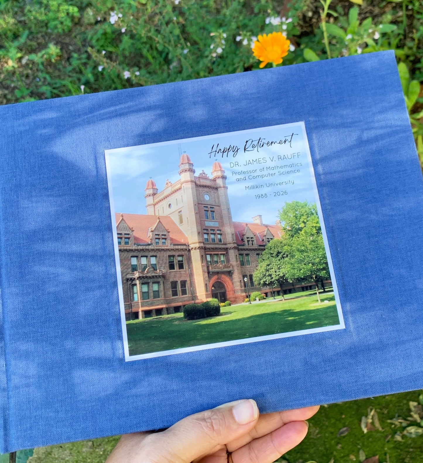 Blue linen retirement party guest book with a photo of a building on the cover held by a hand outdoors.