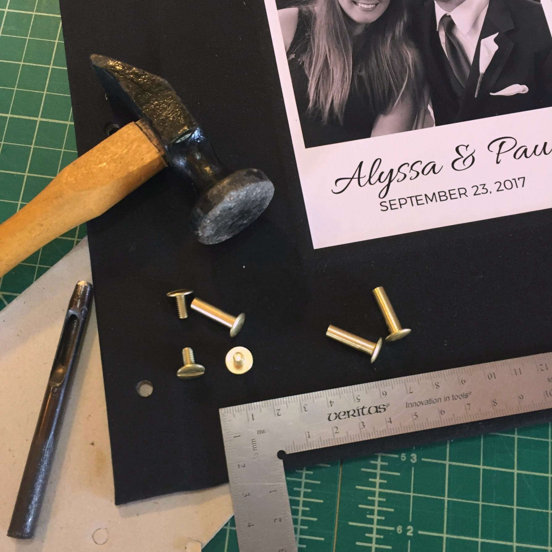 A bindery work table showing a screwpost binder in  process of being made with bookbinding tools: hammer, ruler, brass screw posts, hole punch.