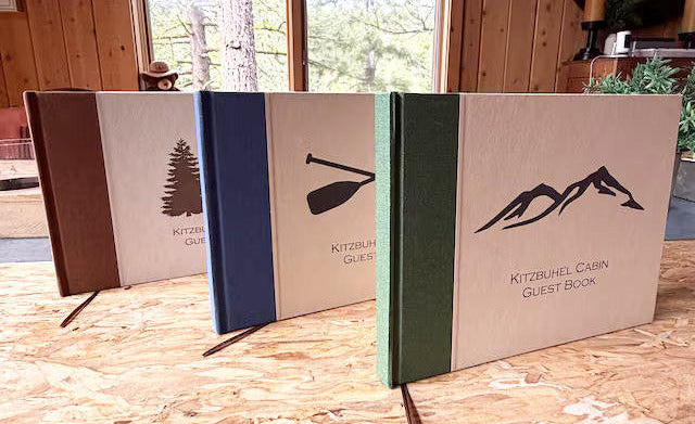 Set of three vacation rental guest books with mountain, tree, and paddle designs on a wooden surface.