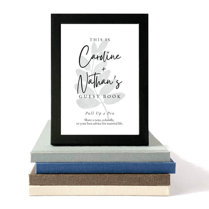Framed guest book sign with decorative elements on a stack of books. Custom table sign for wedding guest made to match personalized hand bound book. Ready for guest book table.