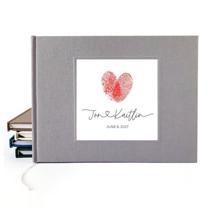 A personalized gray fabric-covered wedding guest book standing upright, with a white insert showing a red fingerprint heart and the names 'Jon & Kaitlin' plus the date 'June 6, 2027.' A stack of books is visible on the left side, and a white ribbon bookmark hangs from the bottom.