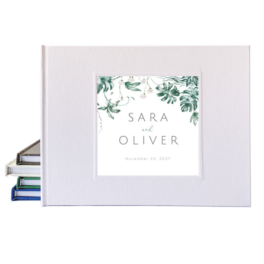 Personalized tropical wedding guest book with a white linen cover, and the bride and groom's names surrounded by tropical leaves.