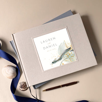 Cream linen wedding guest book with a golden modern minimalist design in cover inlay showcasing bride and groom's name on a beige table with shells, a pen and a blue ribbon.