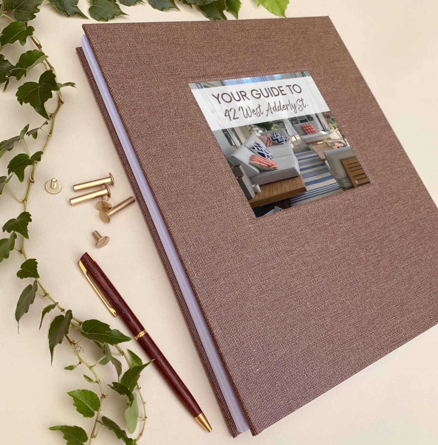 Brown linen welcome binder for airbnb with hard back cover, removable pages displayed on a welcome table with brass screw posts, ivy and a pen. Photo of rental on the cover.