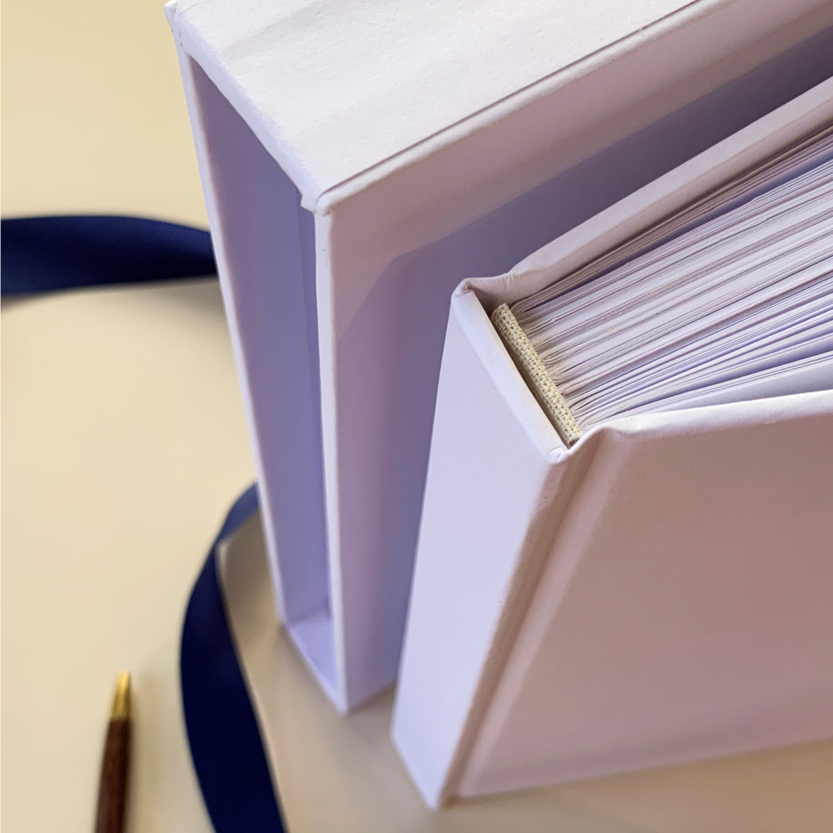 A close-up of a white hardcover  journal standing next to an open matching book slipcase, with a navy ribbon and a small gold pen visible on a light beige surface.